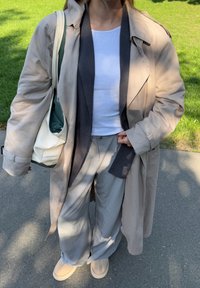 Bežna trenčjak s širokimi rokavi čez temno sivi blejzer in belo rebrasto majico brez rokavov. Svetlo sivi široki hlače in bež natikači.