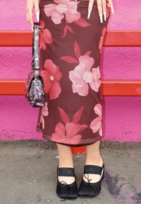 Person, die einen braunen geblümten Rock, schwarze Mary Jane Schuhe trägt und eine kleine gemusterte Handtasche hält, steht vor einer rosa-roten Wand.