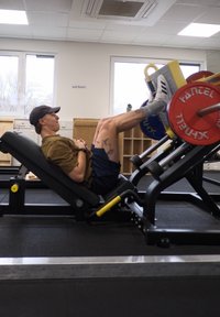 Máquina de press de piernas con discos de pesas de colores. Estructura negra, respaldo inclinado y plataforma para pies ajustable. Entorno de gimnasio con almacenamiento de madera.