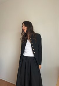 Schwarze, maßgeschneiderte Blazer mit goldenen Knöpfen, getragen über einem weißen Hemd, kombiniert mit einem schwarzen, plissierten Rock. Glatter Stoff und strukturierter Schnitt.