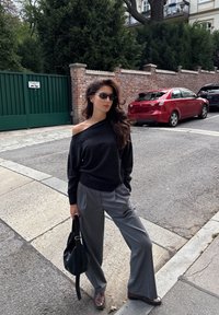 Maglione nero off-shoulder abbinato a pantaloni larghi a righe grigie. Gli accessori includono una borsa nera e occhiali da sole sottili. Scarpe marroni.