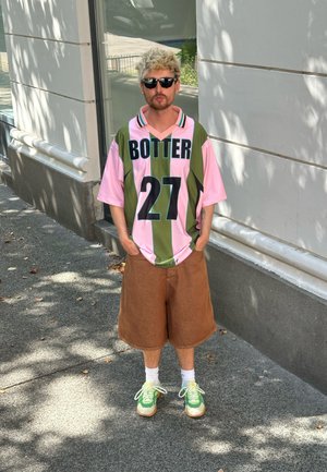Camisa de manga corta de gran tamaño en rayas rosa y oliva con "BOTTER" y "27" impresos; pantalones cortos marrones; zapatillas verdes con acentos amarillos.