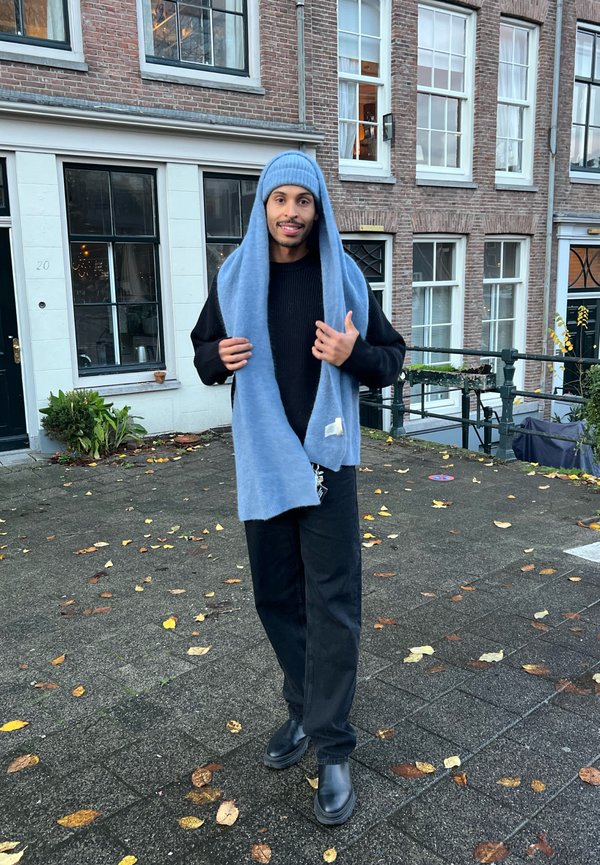 Cachecol de malha azul claro drapeado sobre um suéter preto, combinado com calças pretas e botas pretas, em pé sobre uma calçada texturizada com folhas caídas.