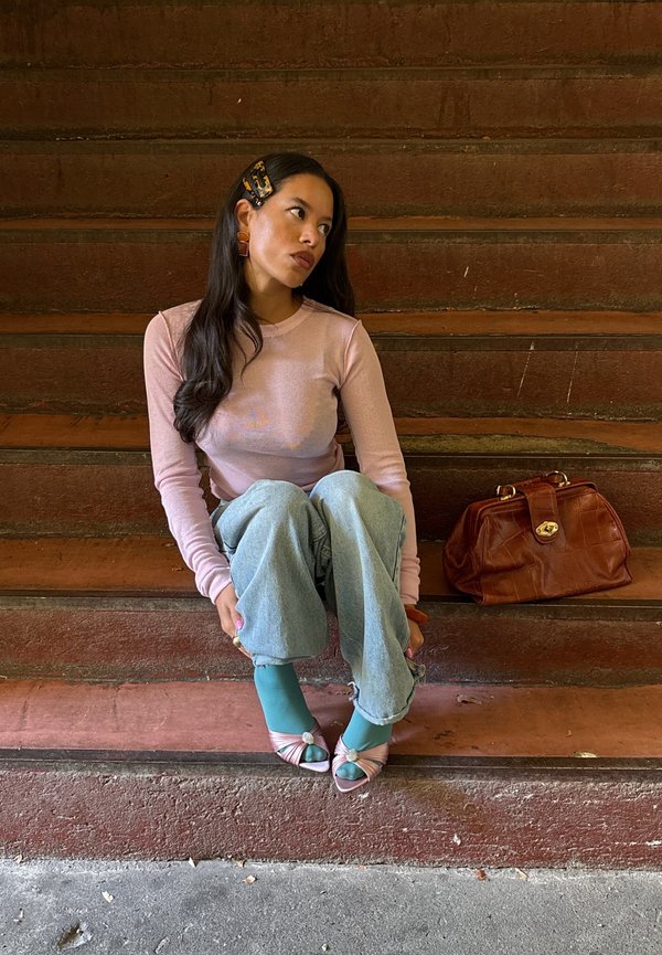 Mujer joven con camiseta rosa de manga larga y vaqueros sentada en escalones de madera marrón con un gran bolso marrón a su lado.