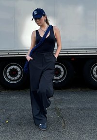 Black bodysuit, loose black trousers, navy blue cap with white logo, textured blue bag, silver jewelry, standing near truck wheels on pavement.