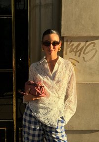 Λευκή δαντελένια μπλούζα με κουμπιά, μπλε και λευκό καρό παντελόνι, ροζ τσάντα χεριού, και μαύρα γυαλιά ηλίου μπροστά σε φόντο από πέτρινο τοίχο.