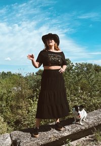 Μαύρο crop top με γραφικό "HERBOLOGY", στρωμένη μαύρη maxi φούστα, μαύρα παπούτσια με ξύλινα τακούνια και μαύρ�ο ψάθινο καπέλο; σκύλος δίπλα.