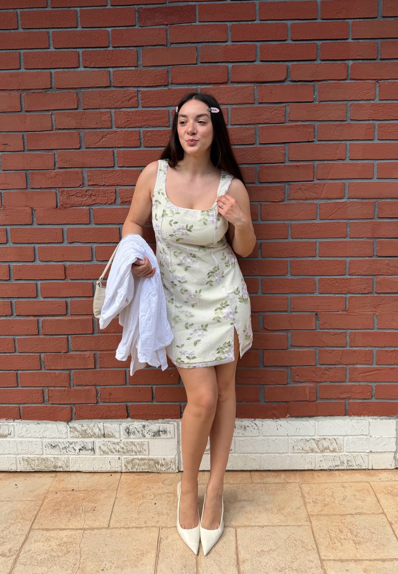 Jeune femme en robe crème à fleurs et talons blancs se tient contre un mur en briques rouges, tenant une veste blanche et un sac beige.