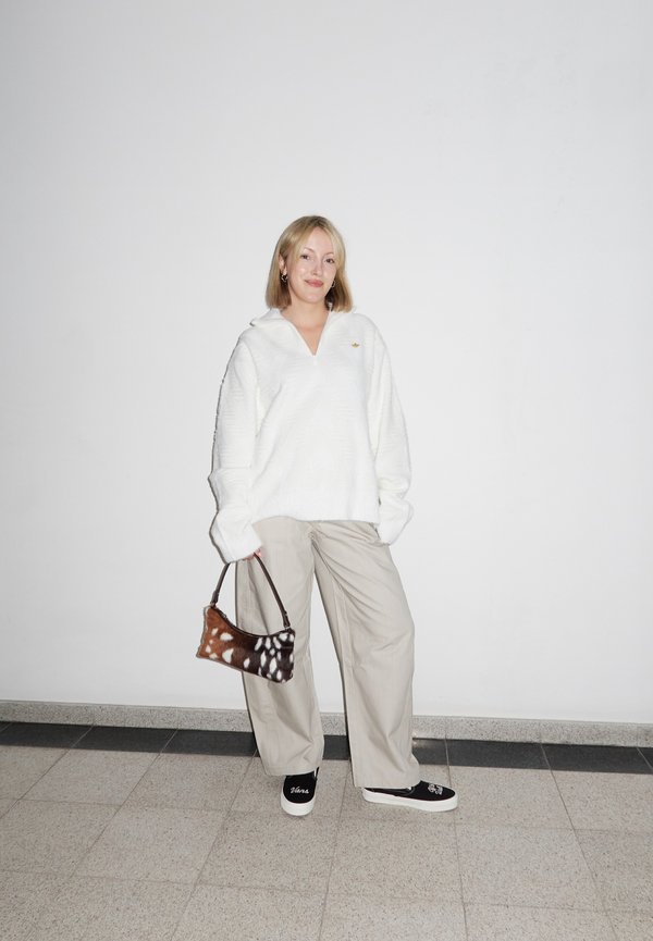 Ung kvinde står mod en hvid væg iført hvid oversized sweater, beige brede bukser, sorte slip-on sko, mens hun holder en brun mønstret håndtaske.