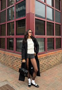 Langer schwarzer Mantel, weißes geripptes Oberteil, graue Shorts, schwarze Strumpfhosen, weiße Socken, schwarze Mary Jane Schuhe, silberne Halsketten, schwarze Handtasche, urbaner Hintergrund.
