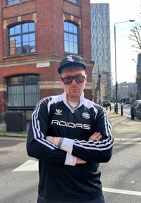 Μαύρη μπλούζα Adidas με μακριά μανίκια και λευκές ρίγες, λογότυπο στο στήθος, συνδυασμένη με σκούρα τζιν και μαύρο καπέλο. Το υπόβαθρο περιλαμβάνει κτίριο από τούβλα.