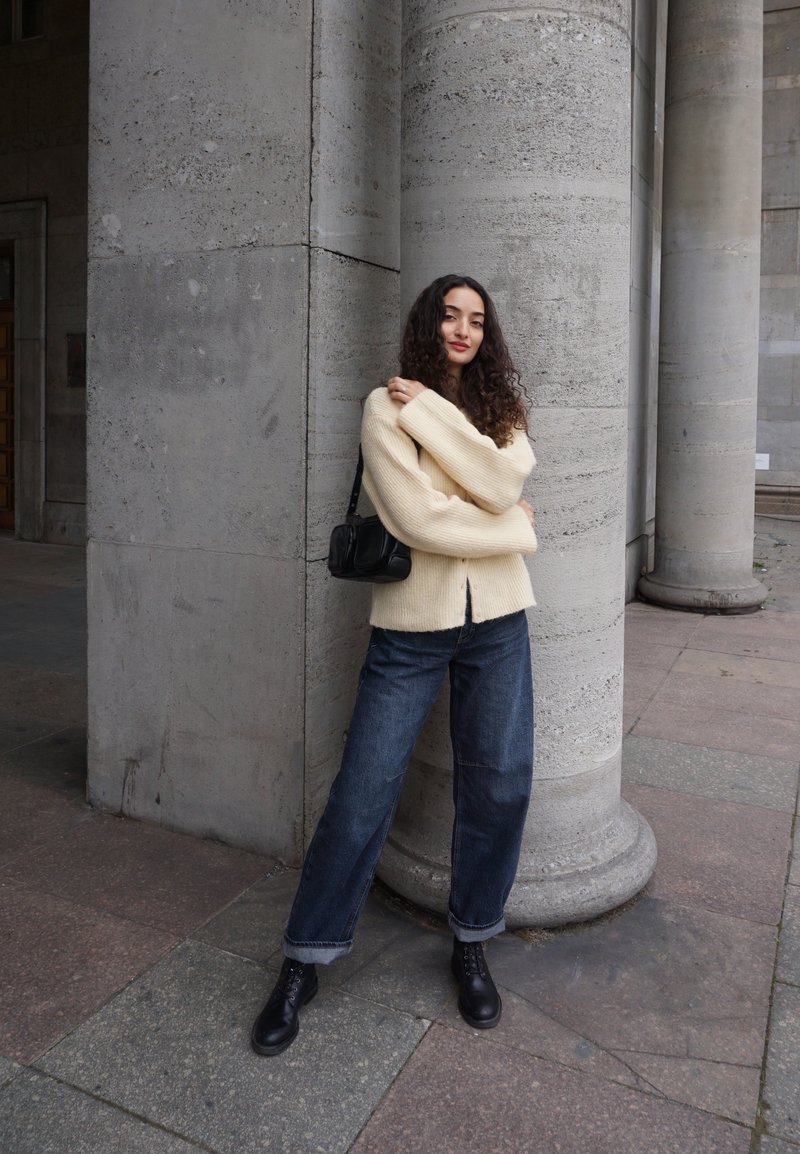Een vrouw met krullend haar draagt een crèmekleurige trui, donkere jeans en zwarte laarzen en leunt tegen een grote stenen pilaar in een buitenomgeving.