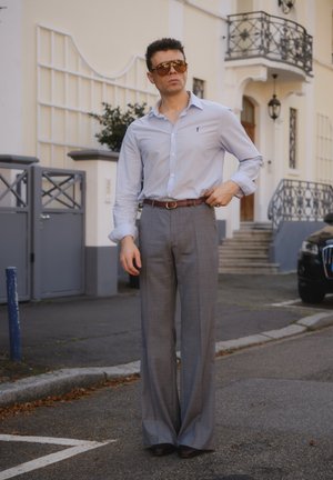 Hellblau gestreiftes Hemd, graue, gefaltete Hose, brauner Ledergürtel mit goldener Schnalle. Das Model trägt große braune Sonnenbrillen. Städtischer Hintergrund.