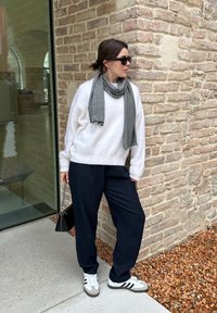 Hvid strikket sweater, grå tørklæde, marineblå bukser og hvide sneakers med sorte og grå detaljer, placeret foran en stenvæg.