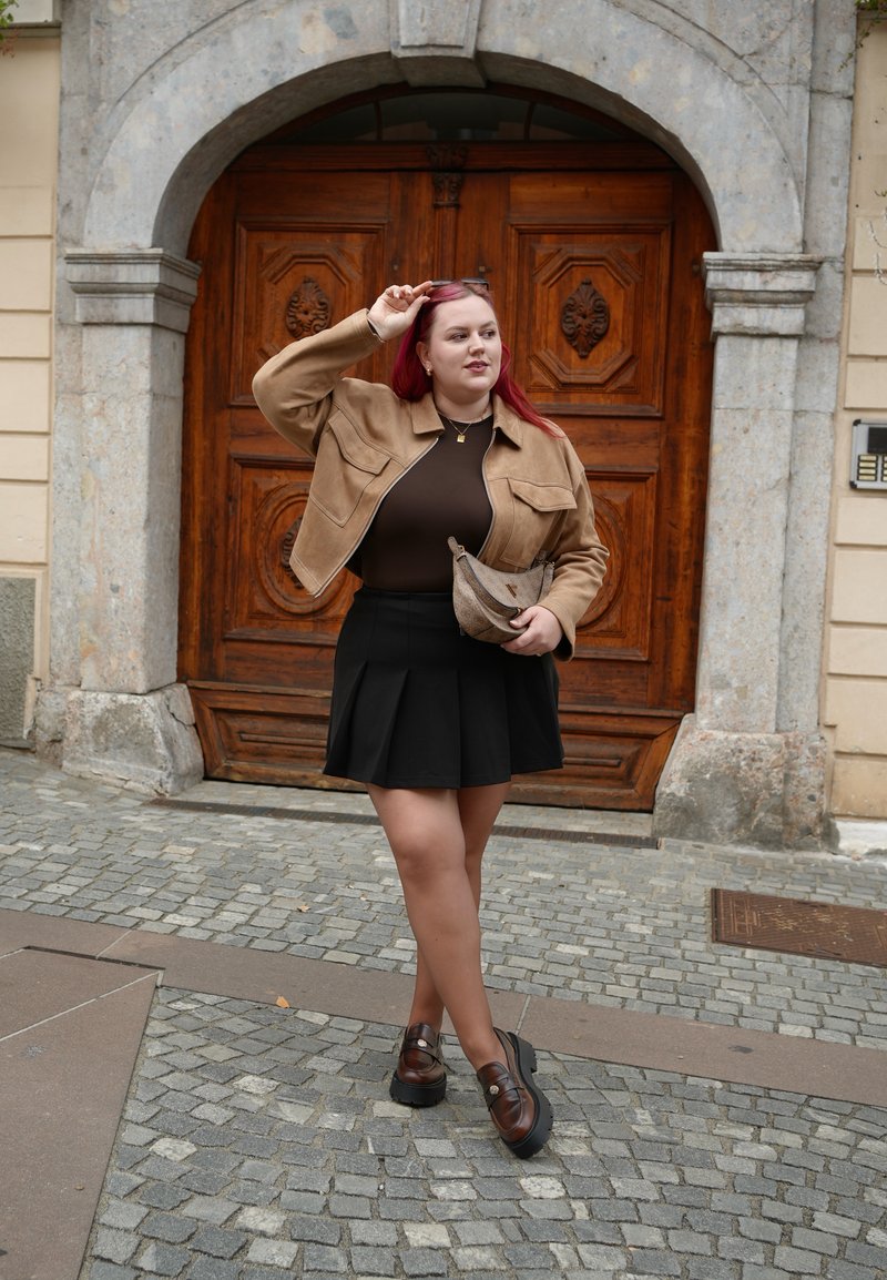 Tan suede cropped jacket, brown top, black pleated skirt, brown and black platform shoes, holding a textured bag, set against wooden doors.