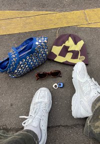 Sac à main bleu clouté, bonnet marron et vert à motif, lunettes de soleil marron, montre de sport blanche, baskets bleu clair à pois sur un trottoir gris.