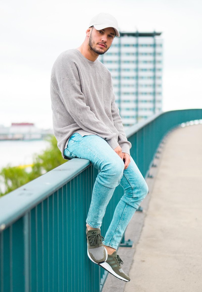 Hellgrauer gestrickter Pullover, hellblaue Slim-Fit-Jeans und olivgrüne Sport Schuhe mit weißen Akzenten, die auf einem grünen Geländer sitzen.