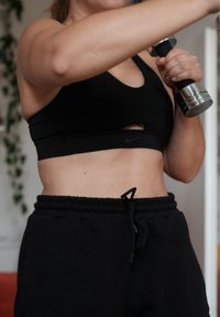 Zwarte sportbeha met uitsnijding, gecombineerd met zwarte trainingsbroeken met een verstelbare trekkoord. Houdt een zilveren dumbbell vast.