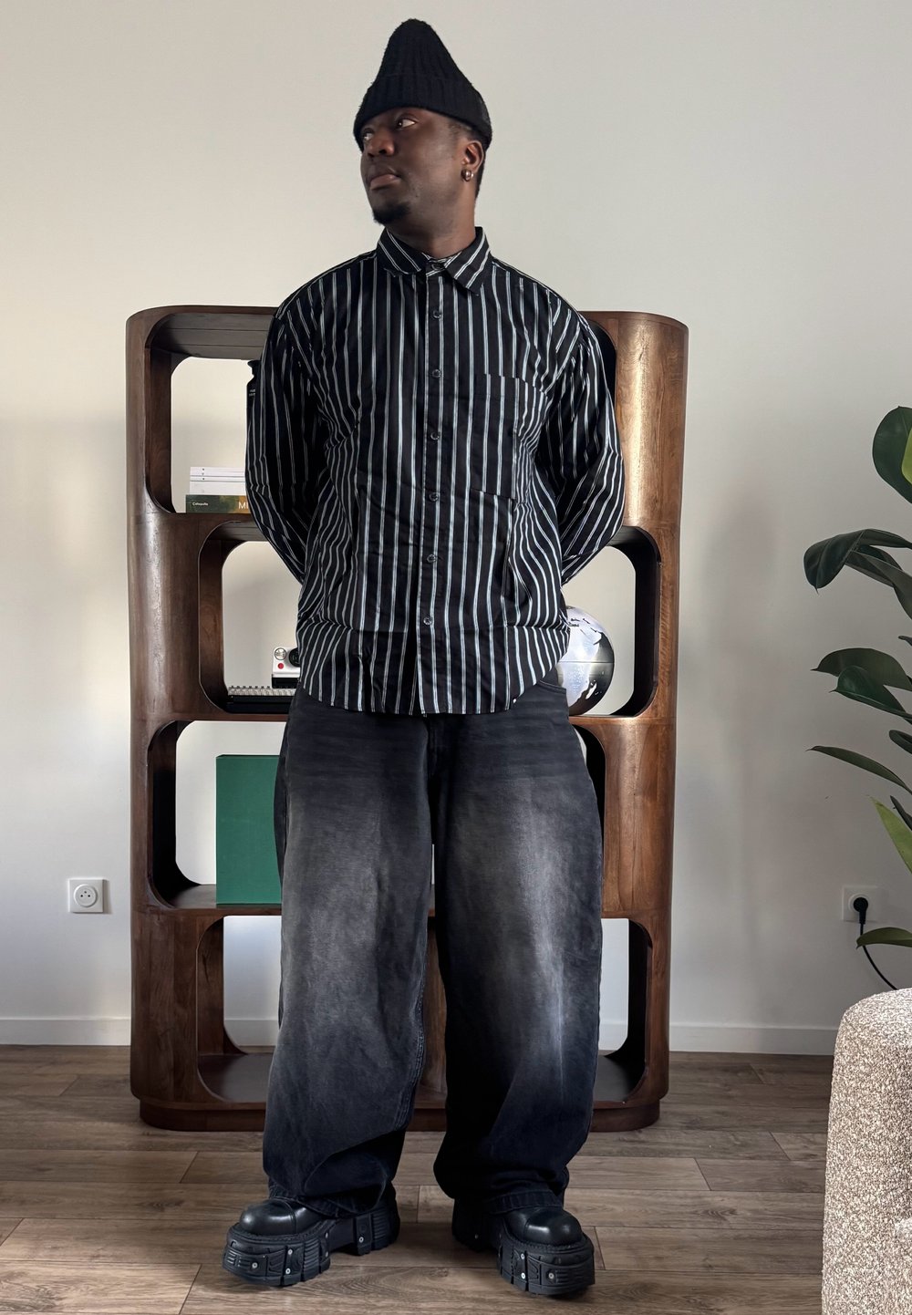 Homme portant un bonnet noir, une chemise rayée noir et blanc, un pantalon large noir et des chaussures noires épaisses, debout à l'intérieur près d'une étagère en bois avec des livres et un globe.