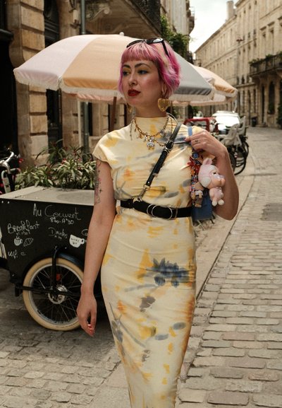 Vestido amarillo con diseño tie-dye, de cuello alto, ceñido al cuerpo y mangas cortas. Los accesorios incluyen joyería dorada y un bolso con charms.