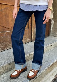 Σκούρες μπλε τζιν παντελόνες με ανοιχτό μπλε τελείωμα, συνδυασμένες με καφέ και λευκά loafers. Το φόντο περιλαμβάνει μια ξύλινη πόρτα και πέτρινες σκάλες.