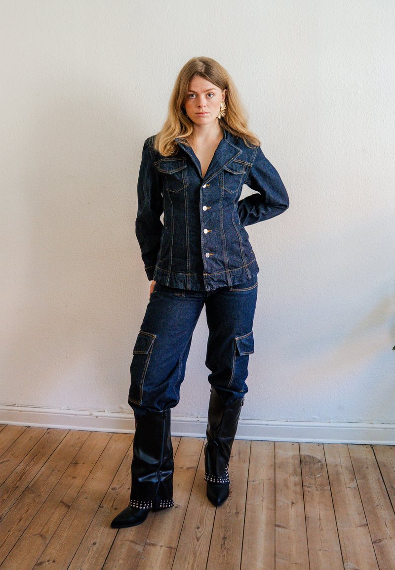 Femme debout à l'intérieur portant une veste et un pantalon en denim foncé avec des bottes noires cloutées montant jusqu'aux genoux, devant un mur blanc et un sol en bois.