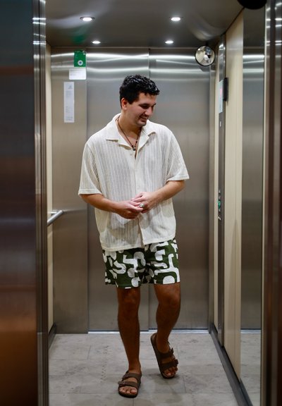 Joven con pantalones cortos estampados y camisa de cuello abierto, de pie en un ascensor de acero inoxidable, mirando hacia abajo y sonriendo, con las manos ligeramente entrelazadas.
