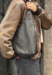 Crna kožna torba za rame s glatkom teksturom, zakrivljenog oblika i uskim remenom, nošena sa strane osobe u smeđoj jakni.