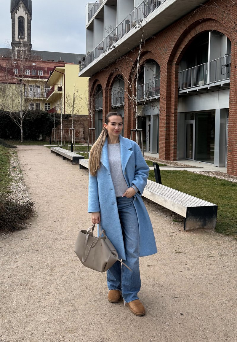 Frau in blauem Mantel und Jeans, die eine beige Tasche hält, steht auf einem Weg nahe modernen Backsteinbögen und Bänken, mit Gebäuden und Bäumen im Hintergrund.