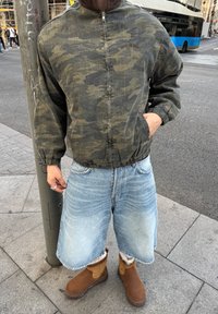 Žmogus stovintis miesto gatvėje, dėvintis kamufliažinę striukę, ilgus džinsinius šortus ir rudus batus su kailiu, viena ranka kišenėje.