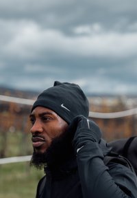 Mann mit Bart, der eine schwarze Mütze, Handschuhe und eine Jacke trägt, passt seine Handschuhe im Freien unter einem bewölkten Himmel an.