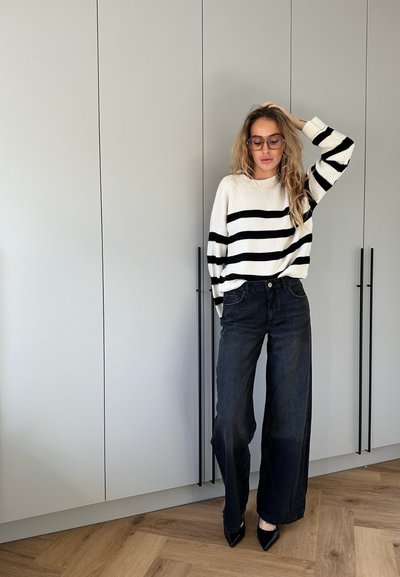 Suéter de punto a rayas en blanco y negro; ajuste holgado. Pantalones de mezclilla de pierna ancha y cintura alta. Tacones negros de punta. Fondo minimalista.