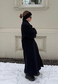 Manteau en laine noire à longue silhouette, porté avec un bandeau noir et des lunettes de soleil, associé à des bottes noires épaisses, debout sur la neige.