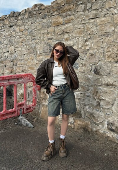 Chaqueta de cuero marrón, camisa blanca ajustada, pantalones cortos de mezclilla, botas marrones con cordones, complementado con gafas de sol. El fondo presenta una pared de piedra y una barricada.