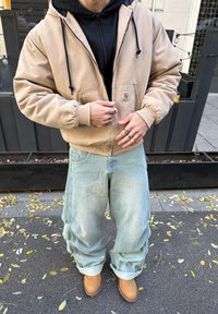 Tan zip-up hoodie met zwarte trekkoorden over een zwarte hoodie, gecombineerd met lichtblauwe baggy jeans en tan laarzen, met een logo op de voorkant van de zak.