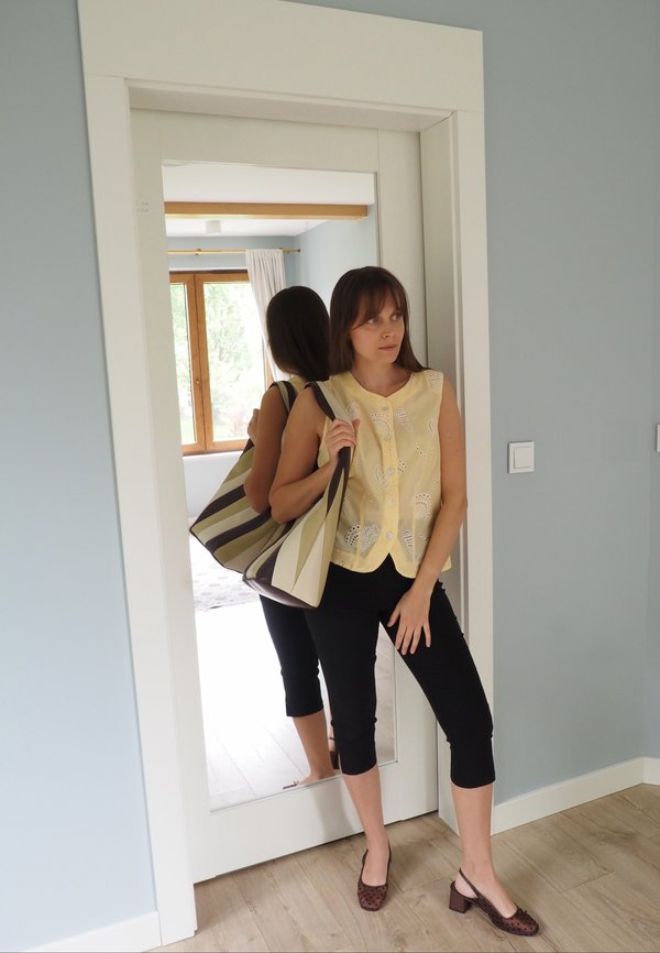 Femme en pantalon noir et haut jaune se tient près d'un miroir en pied tenant un grand sac rayé dans une pièce aux murs bleu clair et au sol en bois.