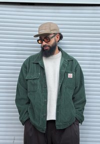 Homme barbu portant une casquette beige, de grandes lunettes teintées, une veste en velours côtelé verte et un pull crème, debout devant un volet métallique argenté.