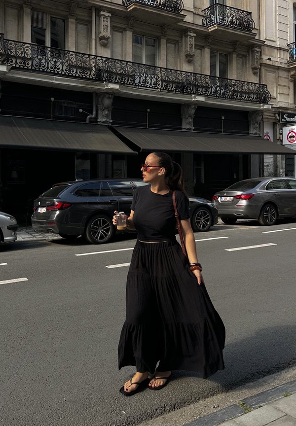 Jupe maxi noire avec un design superposé et une texture fluide, associée à un t-shirt noir cropped et des tongs. Tient une boisson.