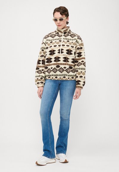 Sudadera de forro polar con cuello alto, base crema, patrones geométricos en marrón oscuro y negro, combinada con jeans acampanados de color azul claro y zapatillas blancas.