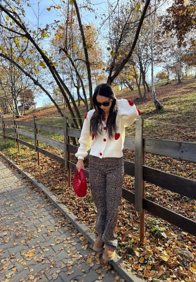 Cárdigan blanco de punto con detalles en forma de corazón rojos, botones rojos, pantalones de estampado leopardo, bolso rojo y zapatos de plataforma beige. Hojas de otoño de fondo.