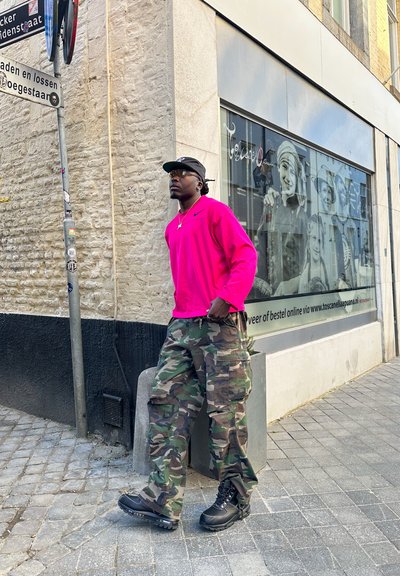 Camisa de manga larga de color rosa brillante, pantalones cargo de camuflaje y botas negras; de pie en una calle de adoquines cerca de una pared texturizada.