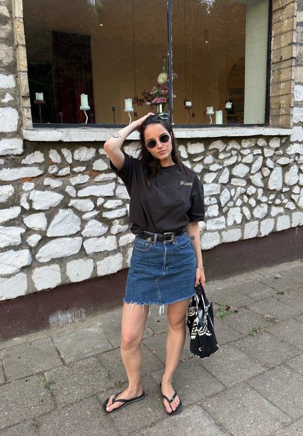 Jeune femme portant des lunettes de soleil, un t-shirt noir, une jupe en jean et des tongs, tenant un sac noir à motifs, debout près d'un mur en pierre avec des bougies dans une fenêtre.