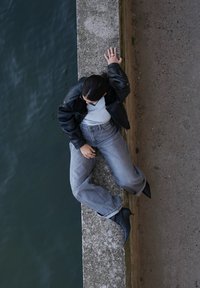 Schwarze Lederjacke, graues T-Shirt, graue weit geschnittene Jeans und schwarze Stiefeletten. Sitzt auf einem Steinrand über Wasser.