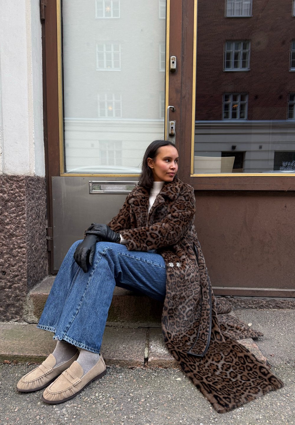 Manteau en fausse fourrure imprimé léopard, pull en maille crème, jean évasé, mocassins beige, gants noirs, assise sur des marches en pierre près d'une porte.