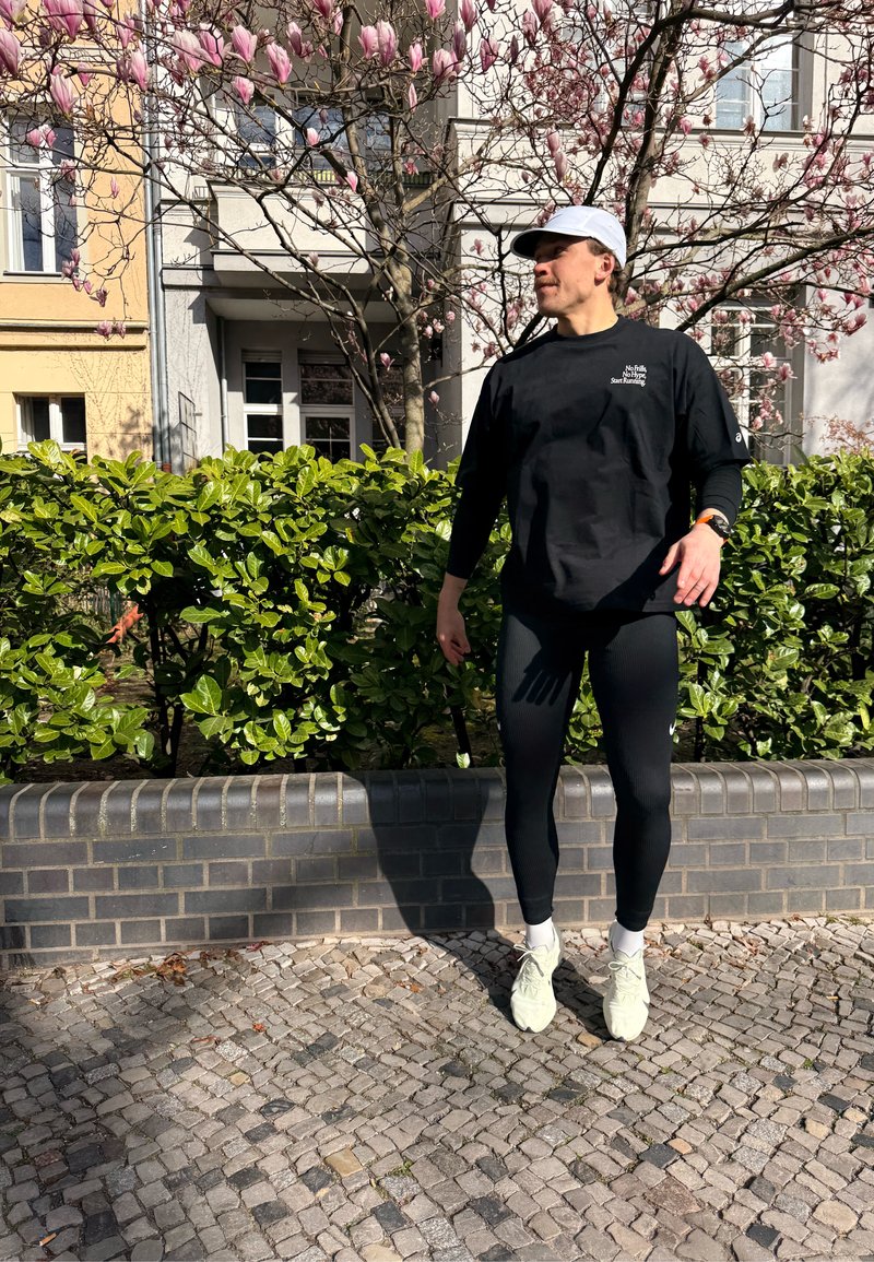 Personne vêtue de vêtements de sport noirs et d'une casquette blanche, debout sur un chemin pavé près de buissons verts et d’un magnolia rose en fleurs, sous la lumière du soleil.