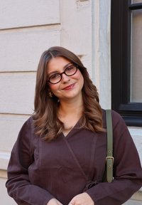 Jeune femme aux cheveux bruns ondulés et portant des lunettes, vêtue d'une robe portefeuille marron foncé et d'un sac à bandoulière vert, souriant près d'un mur en pierre claire.