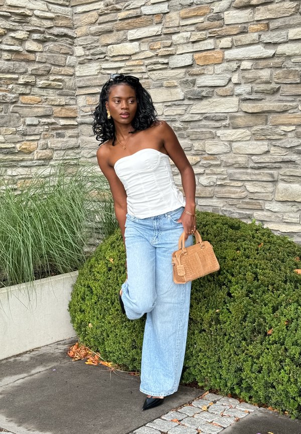 Witte strapless top, lichtblauwe jeans met wijde pijpen, zwarte puntige hakken en een kleine tan handtas met een textuurontwerp. Steenmuur en groen achtergrond.