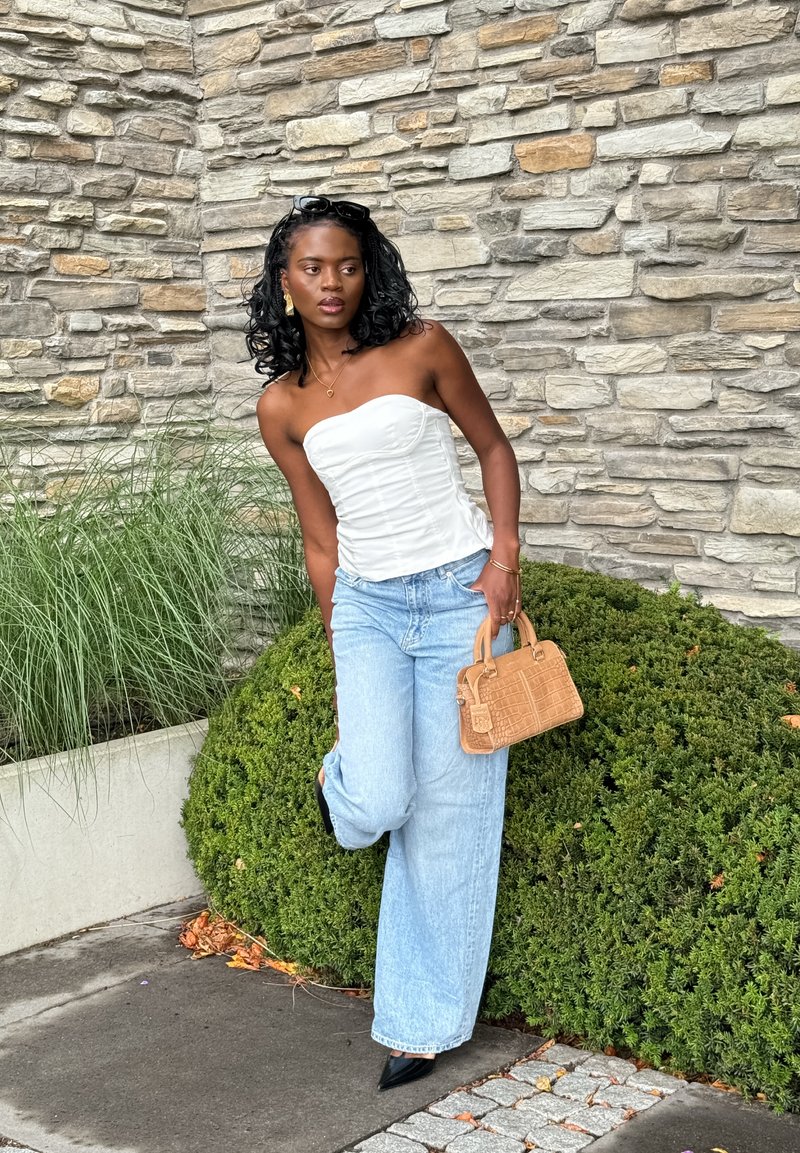 Vit axelbandslös topp, ljusblå vida jeans, svarta spetsiga klackar och en liten beige handväska med texturerad design. Stenvägg och grönskande bakgrund.