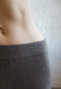 Közeli kép egy mezítelen emberi derékról és köldökről, egy magasított derekú, sötétszürke, bordázott kötött szegély felett.