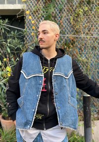 Colete de ganga com bordas desfiadas, quatro bolsos e fechos de botão, sobre um hoodie preto com gráficos coloridos. Ao ar livre, com vegetação.